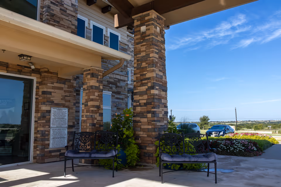 Outdoor seating area at the entrance of a building with stone pillars and two black metal benches with cushions. There are plants and flowers nearby, and cars parked in the background under a clear blue sky.