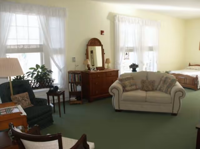 A cozy living space with a green carpet, a white loveseat with patterned pillows, a dark green armchair, a wooden dresser with a mirror and two lamps, a small bookshelf, and a bed in the background near two windows with sheer white curtains.