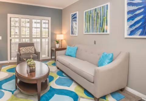 A cozy living room with a light gray sofa adorned with two turquoise pillows, a patterned armchair, a wooden oval coffee table with small potted plants, a colorful abstract rug in shades of blue, green, and gray, three abstract paintings on the wall, and a side table with a lamp and a small plant near a window with white shutters.