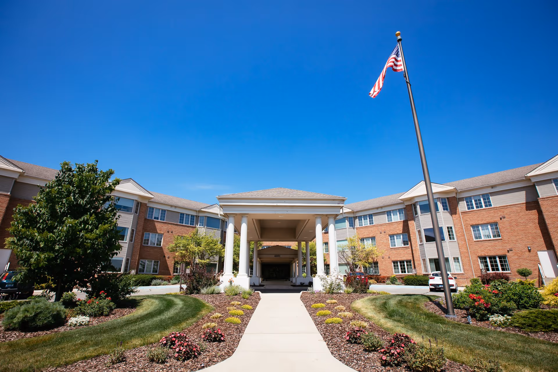 Front exterior view of Glacier Hills Senior Living Community building with a covered entrance supported by white columns, landscaped garden beds with flowers and shrubs, a concrete walkway, an American flag on a tall flagpole, and a clear blue sky.