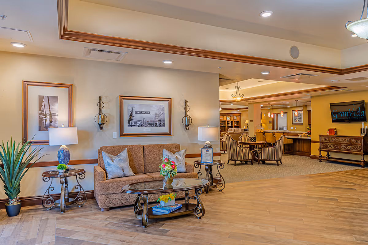 A well-lit senior living facility common area with a brown sofa, glass coffee table with flowers, side tables with lamps, framed black and white photos on the wall, and a large plant. In the background, there is a seating area with chairs around a table, a kitchen or serving counter with pendant lights, and a wall-mounted TV displaying the GranVida Senior Living and Memory Care logo.