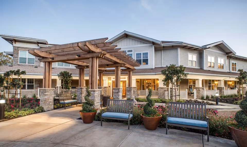 Outdoor seating area with benches and potted plants under a wooden pergola in front of a two-story senior living facility building during early evening.