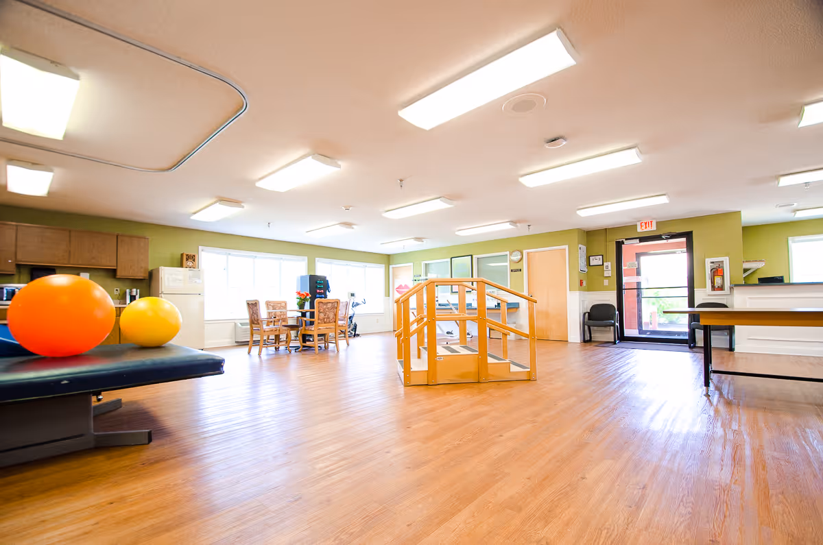 A spacious room with wooden flooring and green walls, featuring a small wooden ramp in the center, a table with chairs near large windows, exercise balls on a padded bench, and a kitchenette area with cabinets and a refrigerator. The room is well-lit with ceiling lights and has an exit door with a glass panel.