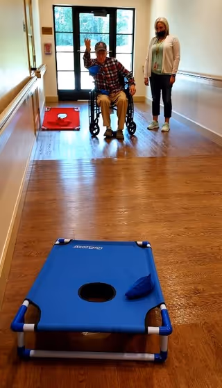 An elderly man in a wheelchair waving while playing a game of cornhole in a hallway with wooden floors. A woman wearing a mask stands nearby watching. There are two cornhole boards set up in the hallway, one blue and one red, with bean bags on them. The hallway has a glass door at the end letting in natural light.