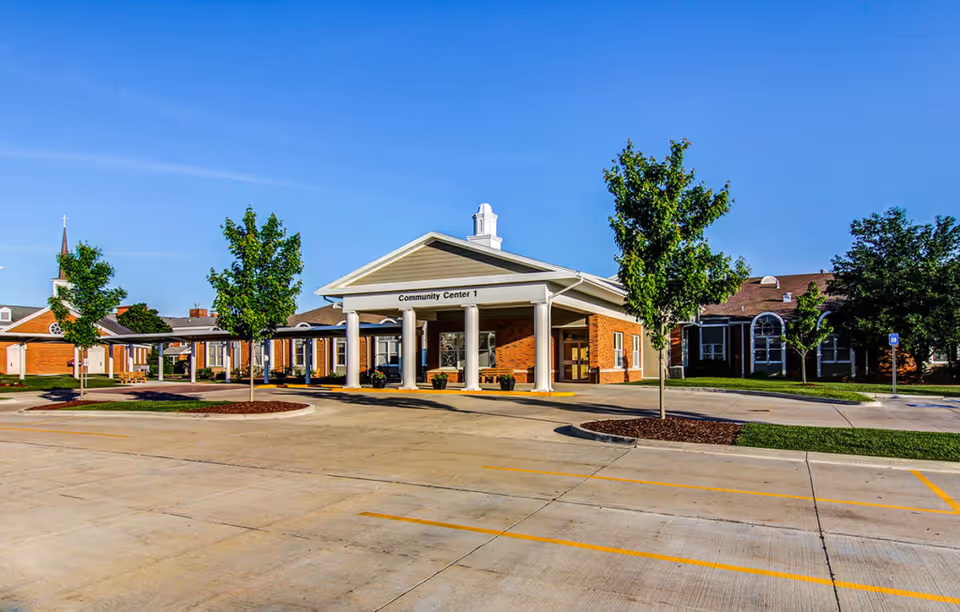 Exterior view of a community center building labeled 'Community Center 1' with a covered entrance supported by white columns, surrounded by trees and a parking lot under a clear blue sky.