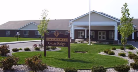 Exterior view of a single-story building with a covered entrance and a flagpole in front. The building is surrounded by well-maintained landscaping with green grass, small bushes, and trees. A sign in front reads 'The Residence at Cedar Ridge.'