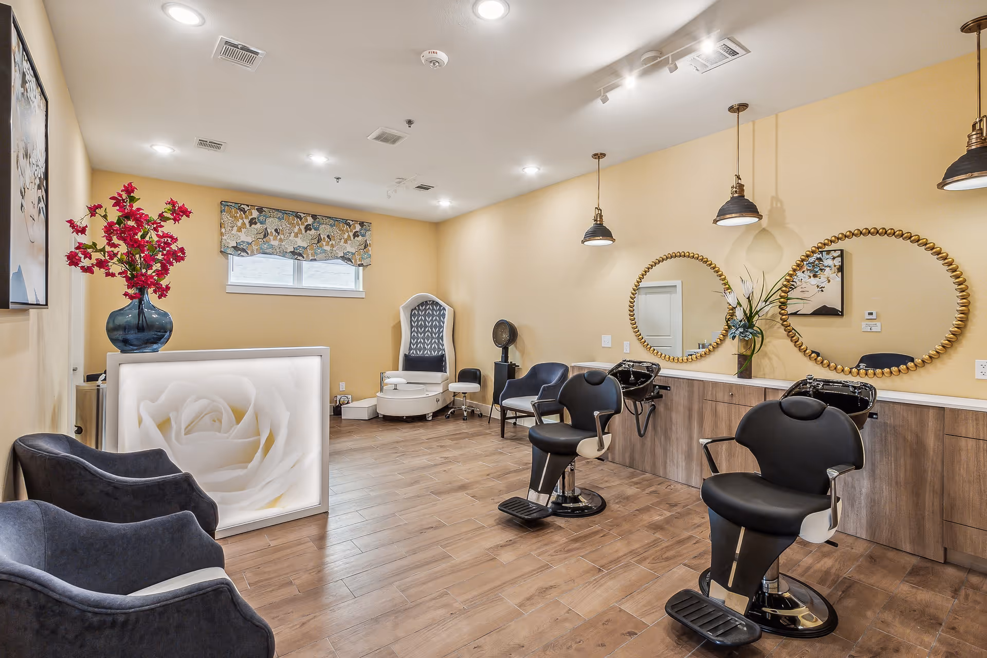 Interior view of a salon area in a senior living facility featuring two black salon chairs with footrests in front of a counter with two round mirrors and wash basins. The room has wooden flooring, beige walls, and ceiling lights. There are two dark blue armchairs on the left side, a reception desk with a large white rose image, a vase with red flowers, and a pedicure chair in the back corner.