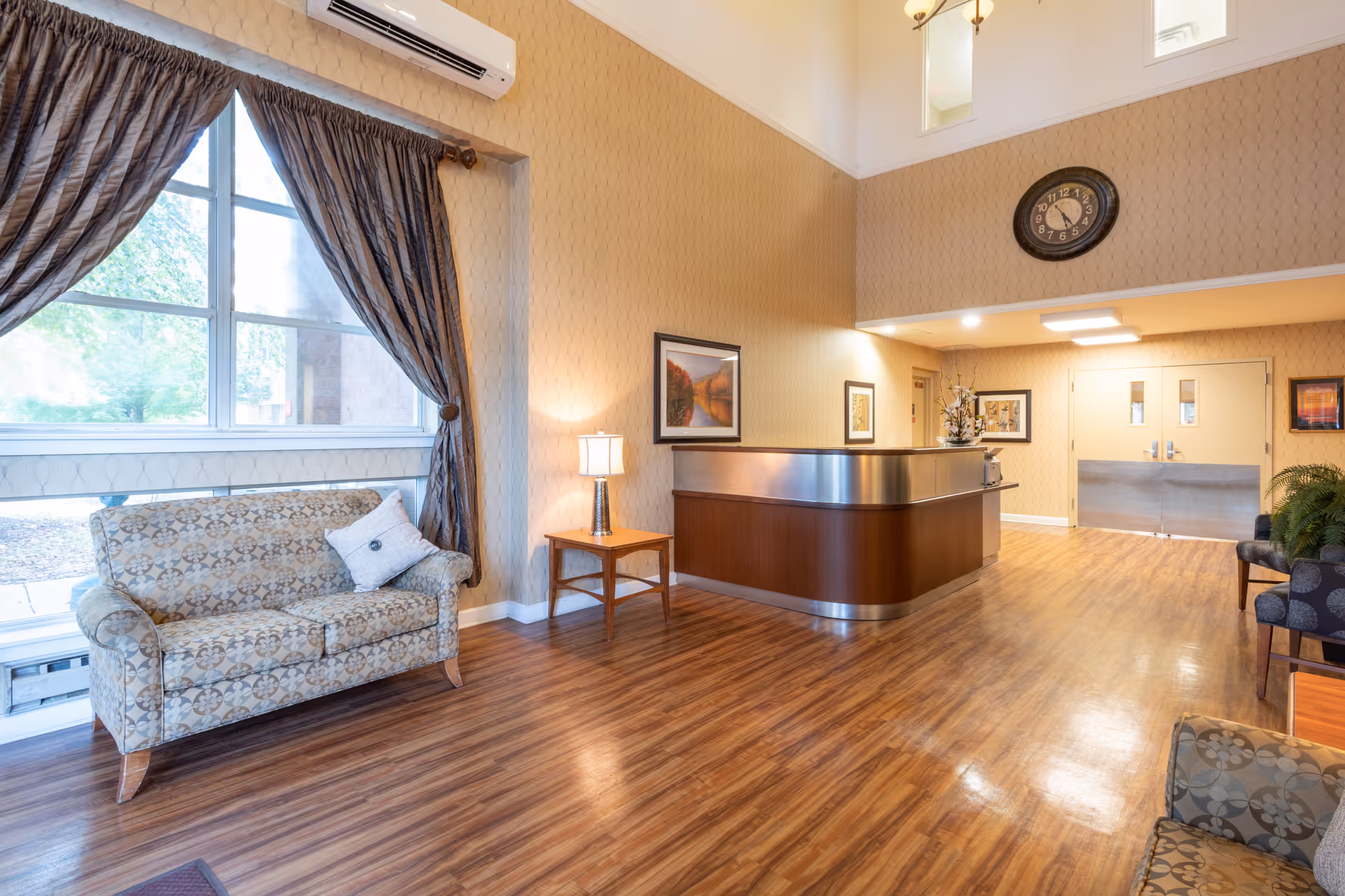 A spacious reception area with wooden flooring, a patterned sofa near a large window with dark curtains, a wooden side table with a lamp, a reception desk with a metallic top, framed artwork on the walls, a large clock above the reception, and double doors in the background.