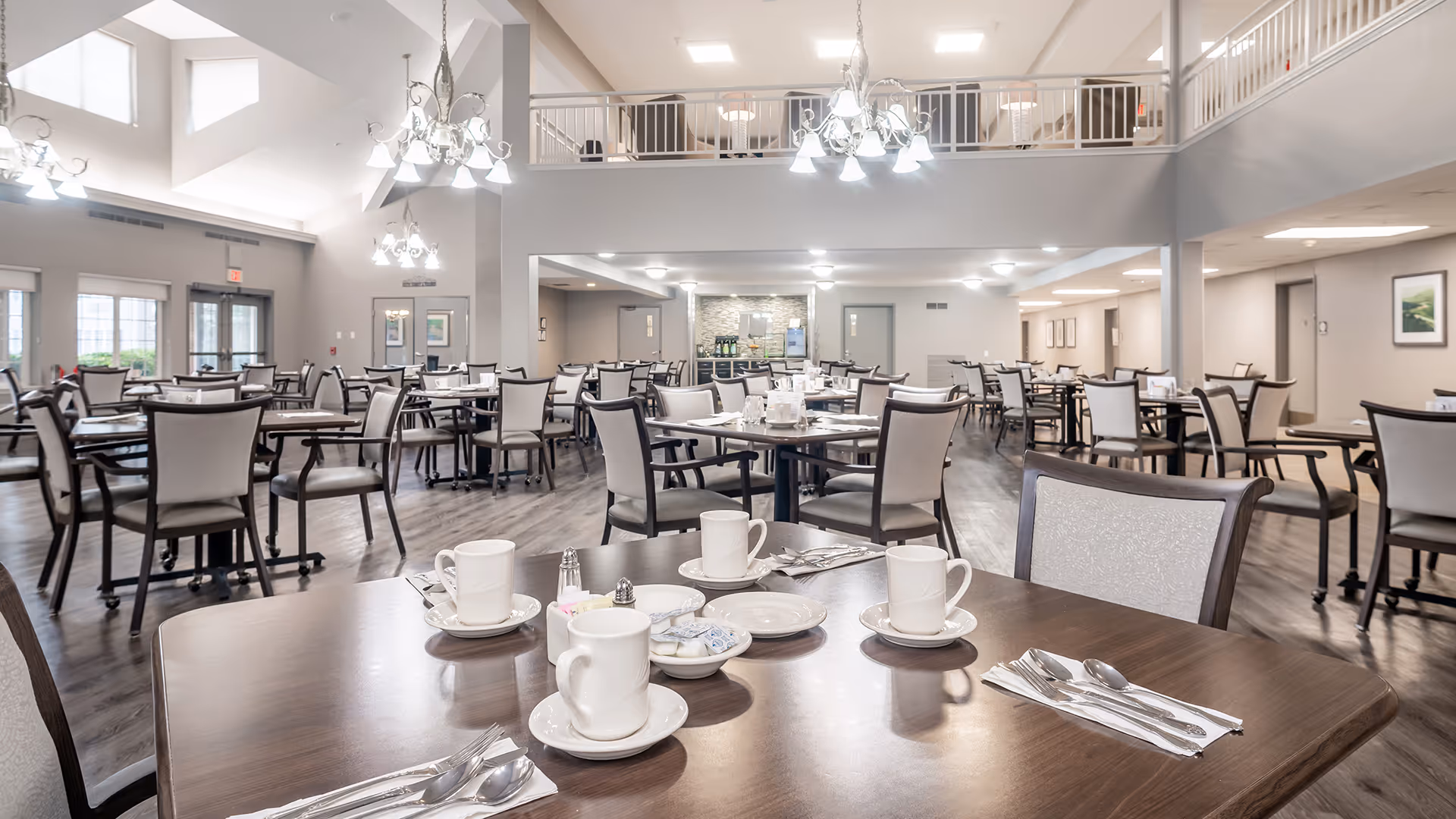 A spacious dining room with multiple tables and chairs arranged neatly. The tables are set with white cups, saucers, plates, and silverware. The room features high ceilings with large windows allowing natural light, modern chandeliers, and a mezzanine level with railings. The flooring is wood, and the walls are painted light gray with framed artwork.