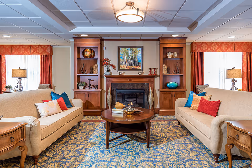 A cozy living room with two beige sofas facing each other, each adorned with colorful throw pillows. Between the sofas is a round wooden coffee table with decorative items on it. Behind the table is a fireplace flanked by two wooden built-in shelves with various decorative objects. The room has patterned blue and gold carpeting, red curtains, and warm lighting from ceiling fixtures and table lamps.