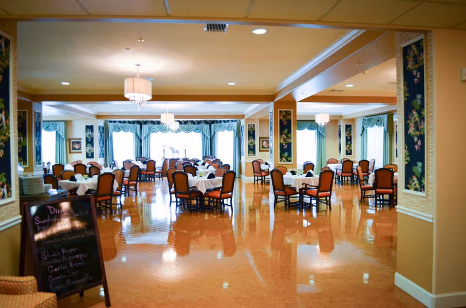 A spacious dining room with multiple round tables covered with white tablecloths, each surrounded by wooden chairs with red cushions. The room features large windows with green curtains and valances, floral wall decorations, and several ceiling light fixtures. A chalkboard menu is visible on the left side near the entrance.
