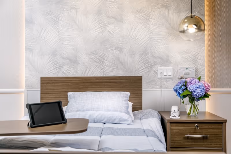 A neatly made single bed with a wooden headboard and gray and white striped bedding. There is a pillow against the headboard. To the right of the bed is a wooden nightstand with a vase of blue and purple hydrangeas, a white telephone, and a drawer. On the left side of the bed is a wooden overbed table with a black tablet on it. The wall behind the bed has a textured light gray and white pattern, with a light switch and a call button panel. A modern round pendant light hangs from the ceiling on the right side.
