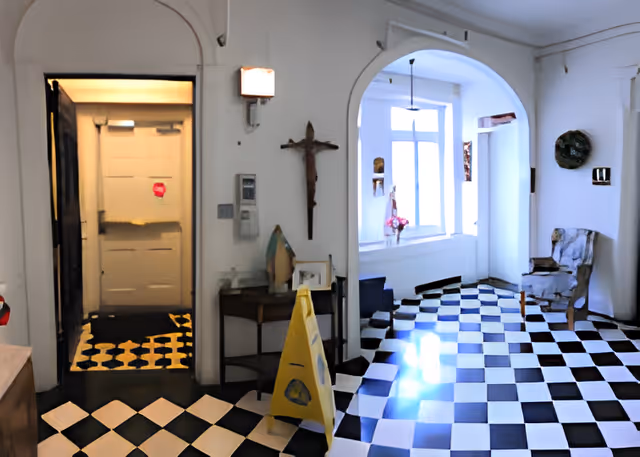 Interior view of a senior living facility room with black and white checkered floor tiles, an arched doorway, a wooden chair with a cushion, a small table with decorative items, a crucifix on the wall, and a large window letting in natural light.