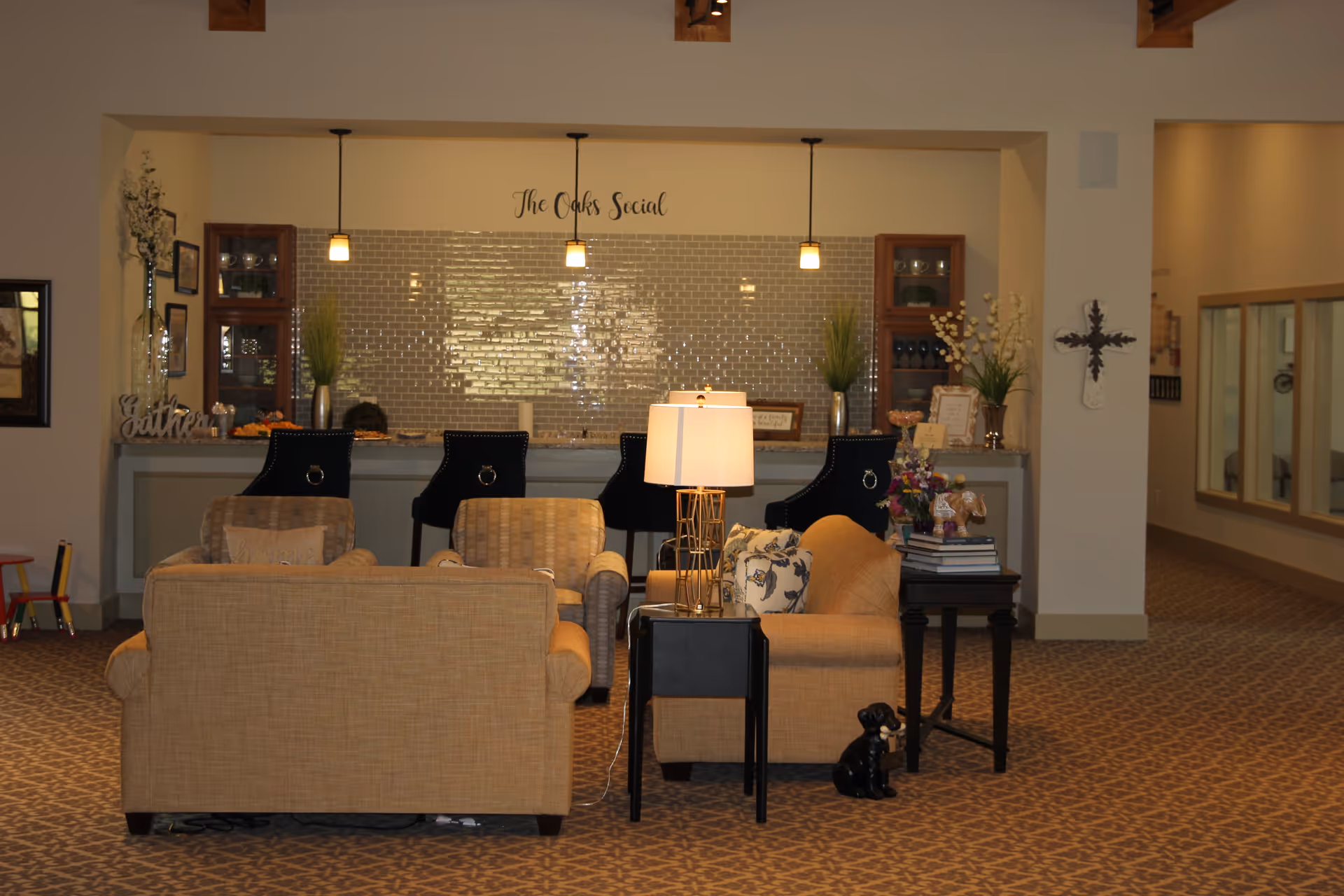 A cozy common area in a senior living facility with beige sofas and armchairs arranged around a small black side table with a lamp. Behind the seating area is a counter with four black bar stools and decorative items, including plants and framed pictures. The wall behind the counter has a tiled backsplash and the words 'The Oaks Social' written above. The carpet has a patterned design, and there is a decorative cross on the wall to the right.