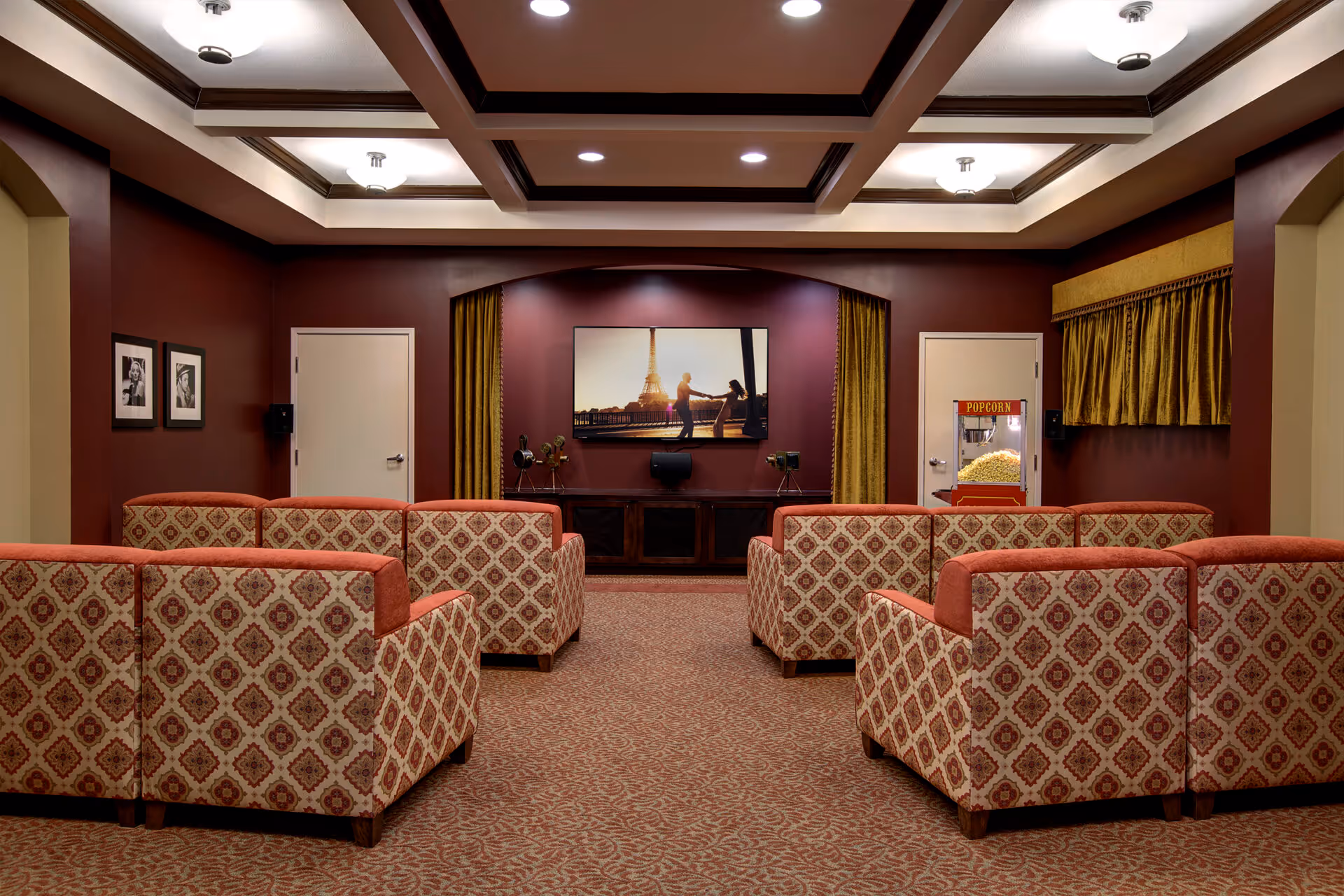 A cozy senior living facility media room with patterned armchairs arranged in rows facing a large flat-screen TV mounted on a burgundy wall. The room features a popcorn machine on the right side, framed black and white photos on the left wall, and a decorative ceiling with recessed lighting.