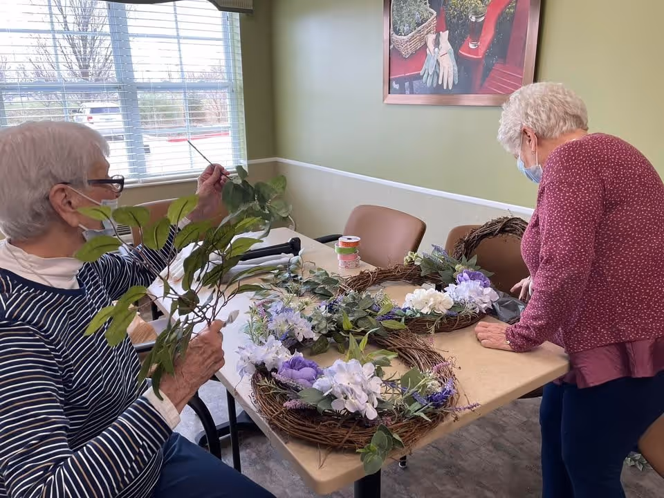 Two elderly women wearing masks are engaged in a craft activity at a table inside a room. One woman is sitting and holding green leaves while the other is standing and leaning over the table. On the table are two decorative wreaths adorned with purple and white flowers, greenery, and craft supplies. The room has a window with blinds, green and white walls, and a framed picture of gardening gloves and plants on the wall.