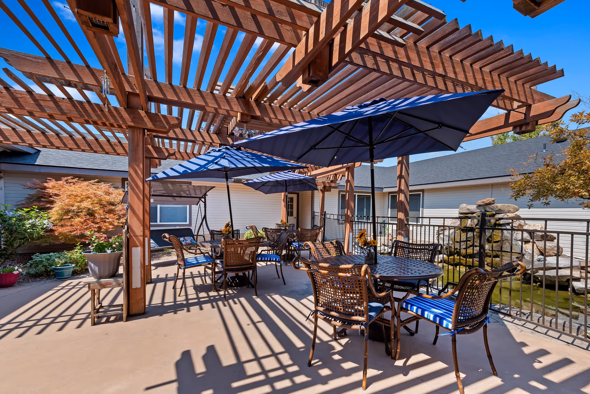 Outdoor patio area at Autumn Wind Assisted Living featuring several round tables with wicker chairs and blue umbrellas under a wooden pergola. There are potted plants and a small water feature with rocks in the background, along with a clear blue sky.