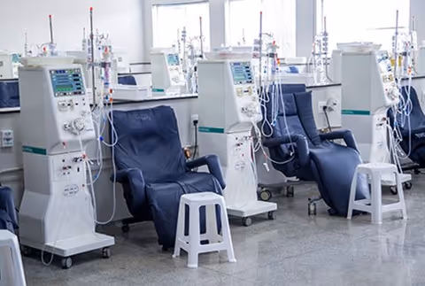 A row of dialysis machines next to reclining chairs with small white stools in front of each chair in a clean, bright medical treatment room.