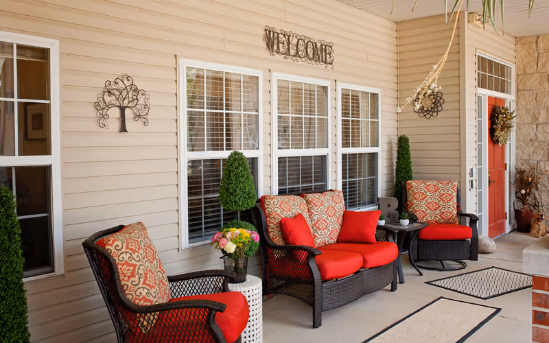 A cozy outdoor porch area with wicker furniture featuring red and patterned cushions. There are three chairs and a loveseat arranged around small tables with decorative plants and flowers. The beige siding wall has three windows with white blinds and a metal 'WELCOME' sign above them. The porch floor has several beige and black rugs, and a red door with a wreath is visible at the far end.