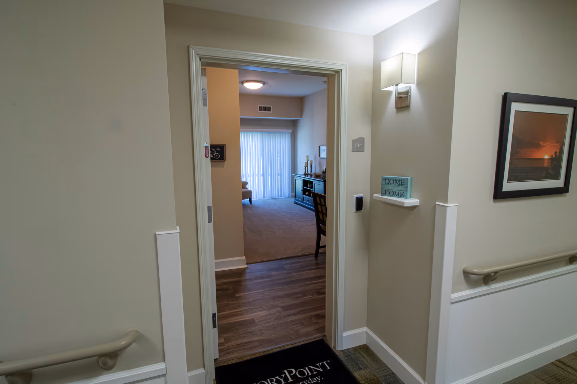 View through an open doorway from a hallway into a living room area with carpeted floor, a chair, a cabinet with decorative items, and a large window with vertical blinds. The hallway has beige walls with handrails, a framed picture, a wall-mounted light fixture, and a small shelf with a decorative sign that says 'HOME'.