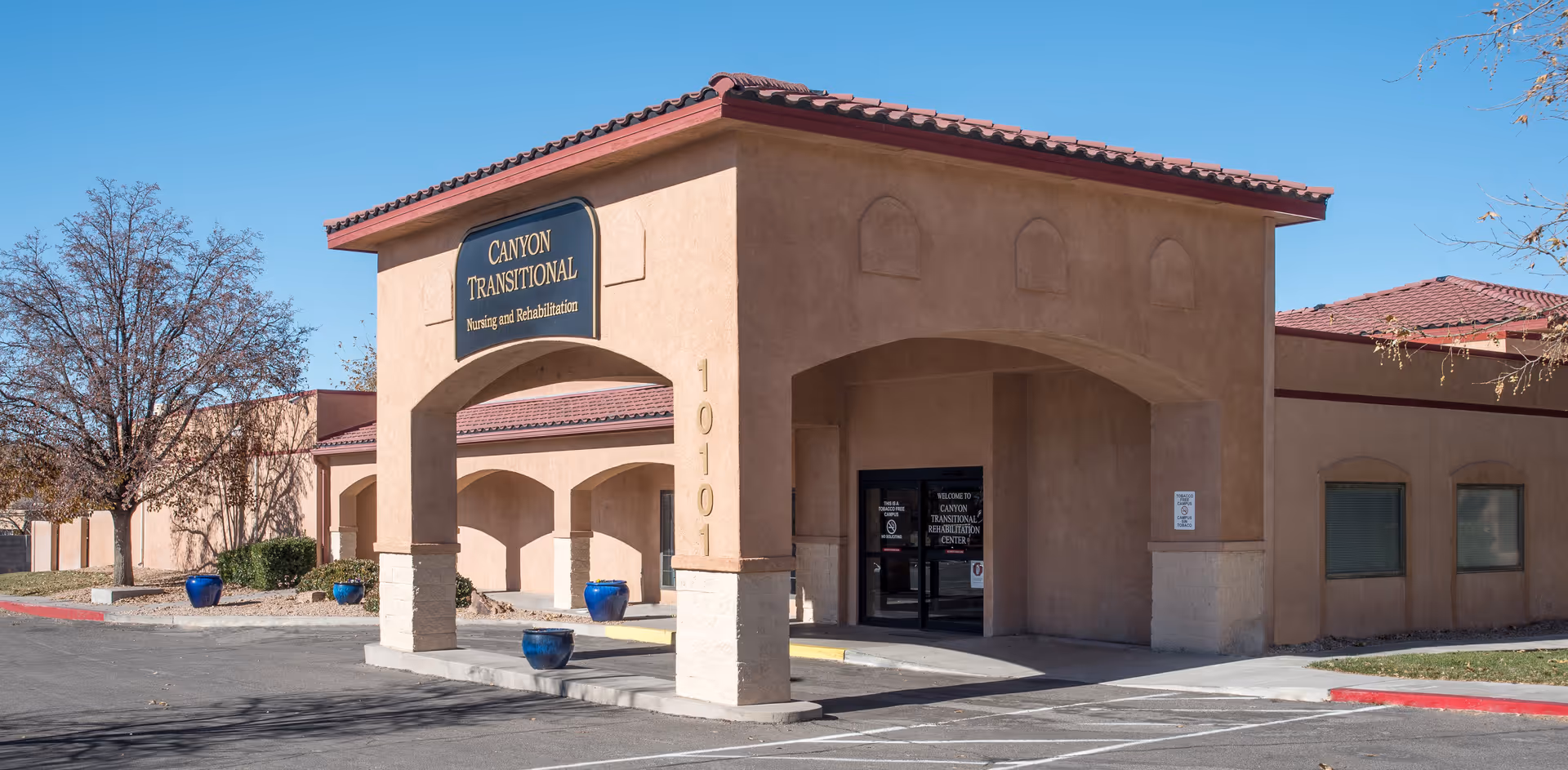 Exterior view of Canyon Transitional Rehabilitation Center building with a covered entrance, beige stucco walls, and a red tile roof under a clear blue sky. There are a few leafless trees and blue planters near the entrance.