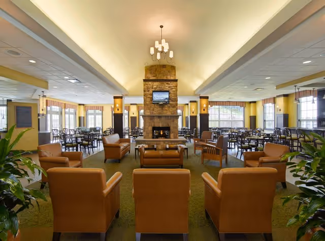 Spacious senior living facility common area with multiple brown leather armchairs arranged around a central stone fireplace with a mounted TV above it. The room has large windows allowing natural light, several dining tables and chairs in the background, green carpet, yellow walls, and ceiling lights including a chandelier above the fireplace.