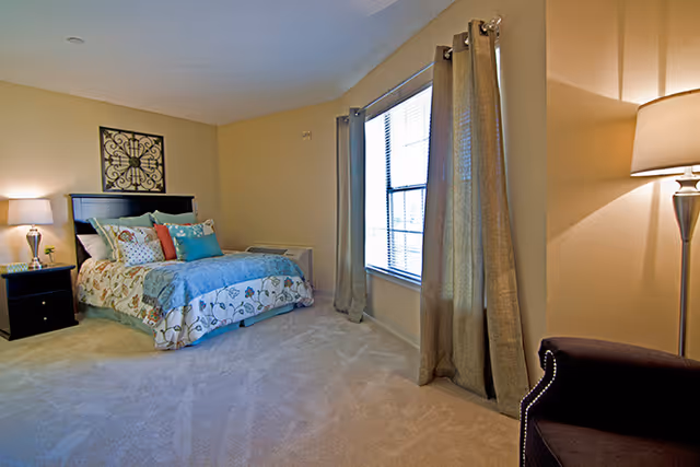 Bright furnished bedroom with a bed, nightstands and lamps, a large window with curtains, and an upholstered chair.