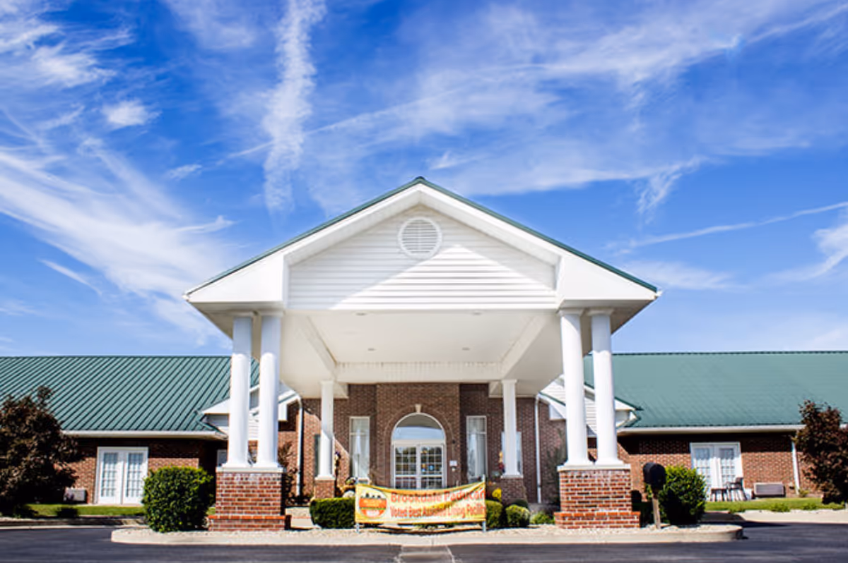 Front entrance of a one-story brick senior living building with white columns and a green metal roof under a blue sky.