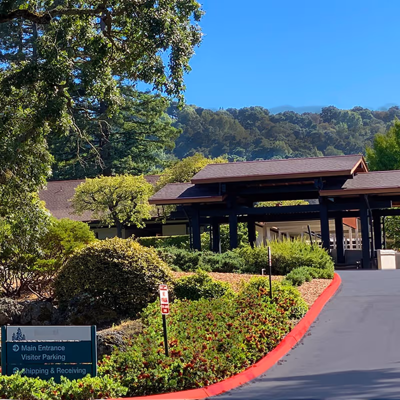 Exterior view of The Sequoias Portola Valley facility entrance with a covered driveway, surrounded by green bushes, trees, and hills in the background under a clear blue sky.
