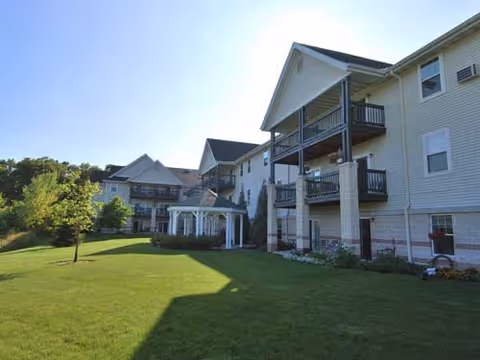 Exterior view of a multi-story senior living facility with beige siding, balconies, and a white gazebo on a well-maintained green lawn under a clear blue sky.