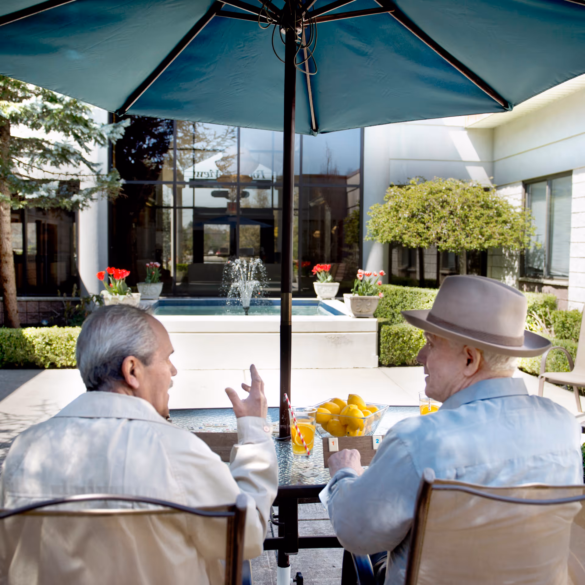 Two elderly men sitting at a glass table under a large blue umbrella in an outdoor courtyard. They are engaged in conversation, with a small fountain and potted flowers visible in the background. One man is wearing a light-colored jacket, and the other is wearing a light blue shirt and a beige hat.