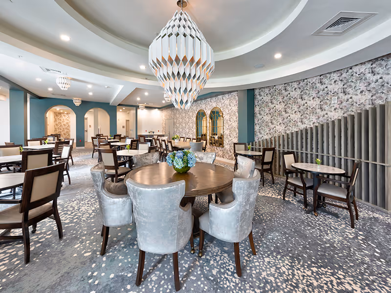 A spacious, elegantly decorated dining room with multiple round and rectangular tables surrounded by chairs. The room features a large, modern chandelier hanging from a circular recessed ceiling. The walls have floral wallpaper and decorative mirrors, and the carpet has a patterned design.