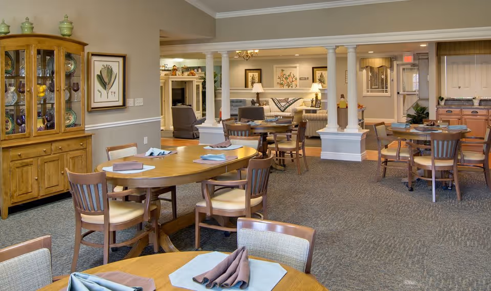 A spacious senior living dining area with multiple wooden tables and chairs set with folded napkins and placemats. The room features a wooden china cabinet with glassware, framed botanical artwork on the walls, and a cozy living area with armchairs and a sofa in the background. The space is warmly lit with lamps and ceiling lights, and has carpeted flooring.
