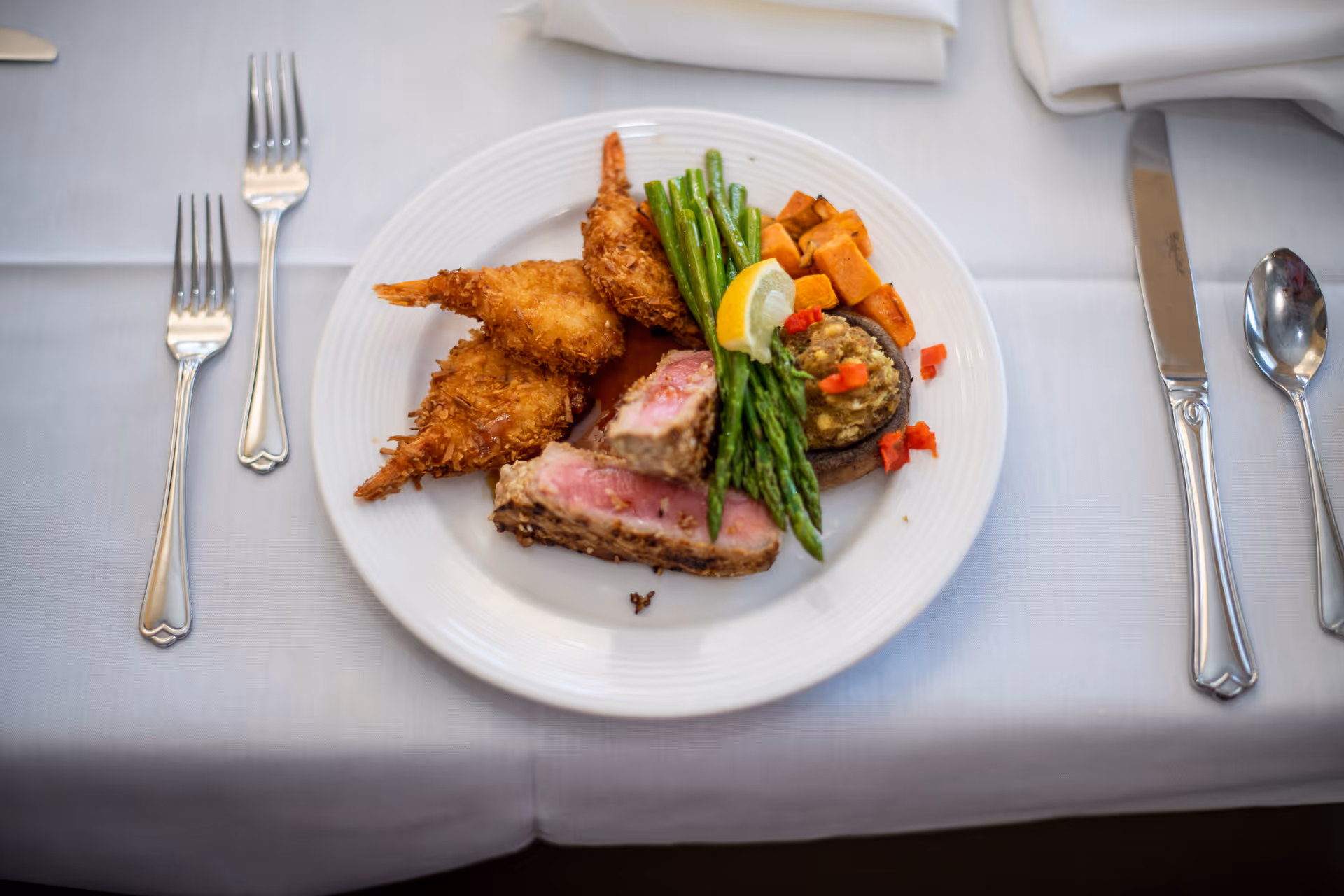 A white plate on a white tablecloth with a meal consisting of two breaded fried shrimp, green beans, roasted sweet potatoes, a stuffed mushroom topped with diced red peppers and a lemon wedge, and two slices of rare cooked meat. The plate is flanked by two forks on the left and a knife and spoon on the right, with a folded white napkin above the knife and spoon.