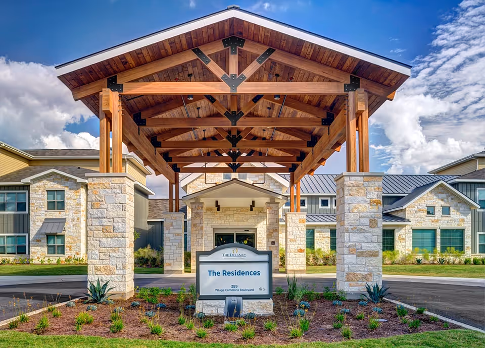 Front entrance of The Delaney at Georgetown Village featuring a large wooden pergola supported by stone pillars, with a sign that reads 'The Residences, 359 Village Commons Boulevard' in front of a landscaped area and residential buildings in the background under a partly cloudy sky.