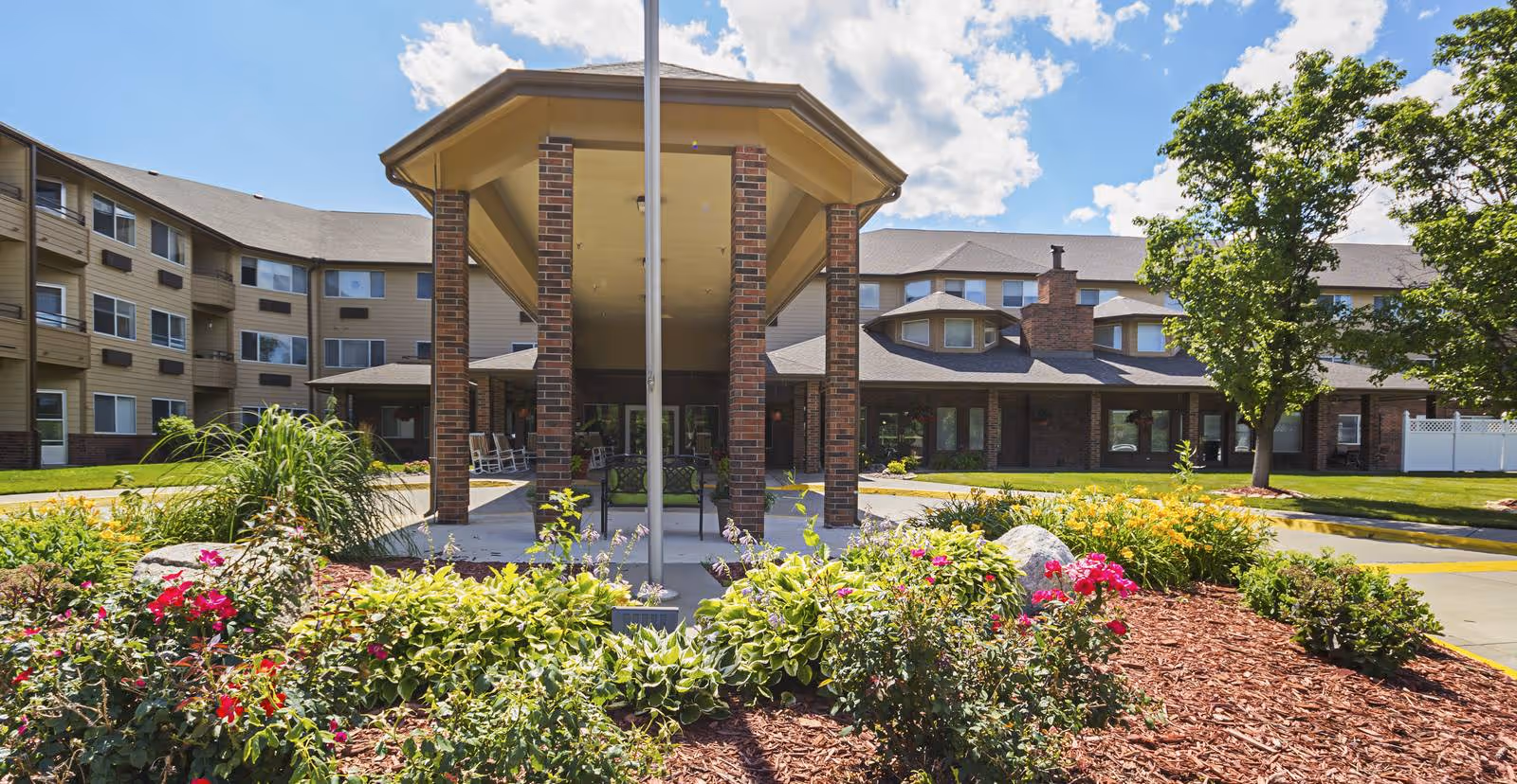 Front exterior view of Holiday Country Squire facility with a covered entrance supported by brick pillars, surrounded by landscaped flower beds and greenery under a partly cloudy blue sky.