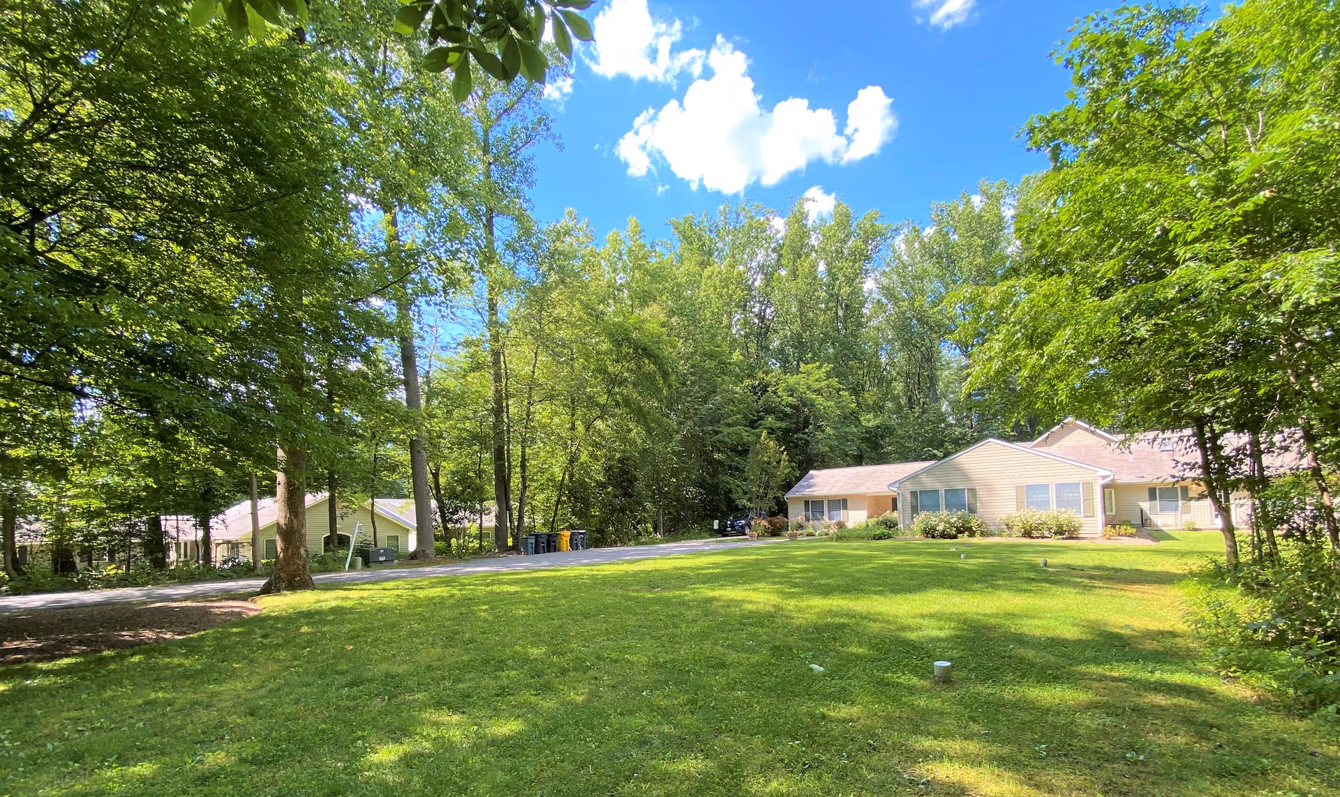 A sunny outdoor scene at Arcadia Assisted Living of Crofton featuring a large green lawn, several trees, and a beige single-story building with multiple windows. The sky is bright blue with a few white clouds.