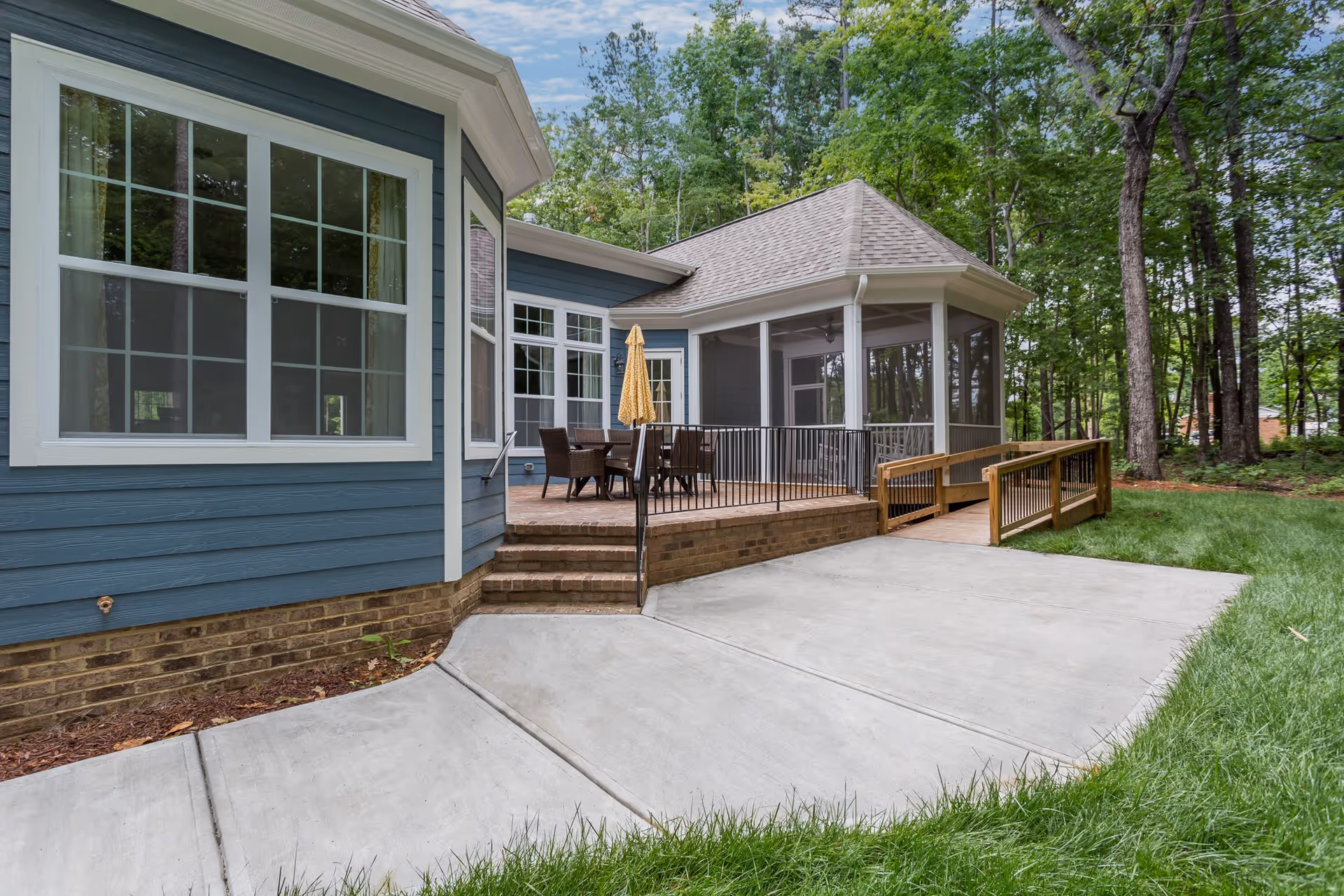 Outdoor patio area of Avendelle Assisted Living on Maynard with a blue exterior building, large windows, a brick patio with a table and chairs under a yellow umbrella, a ramp for accessibility, surrounded by green grass and trees.