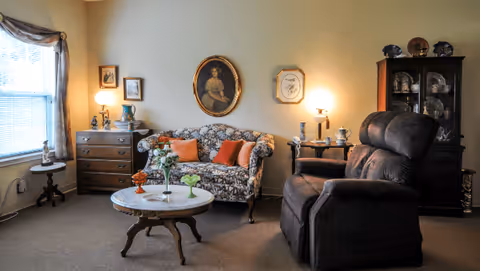 Cozy living room with a floral loveseat, recliner, round coffee table, and antique cabinets under warm lighting.