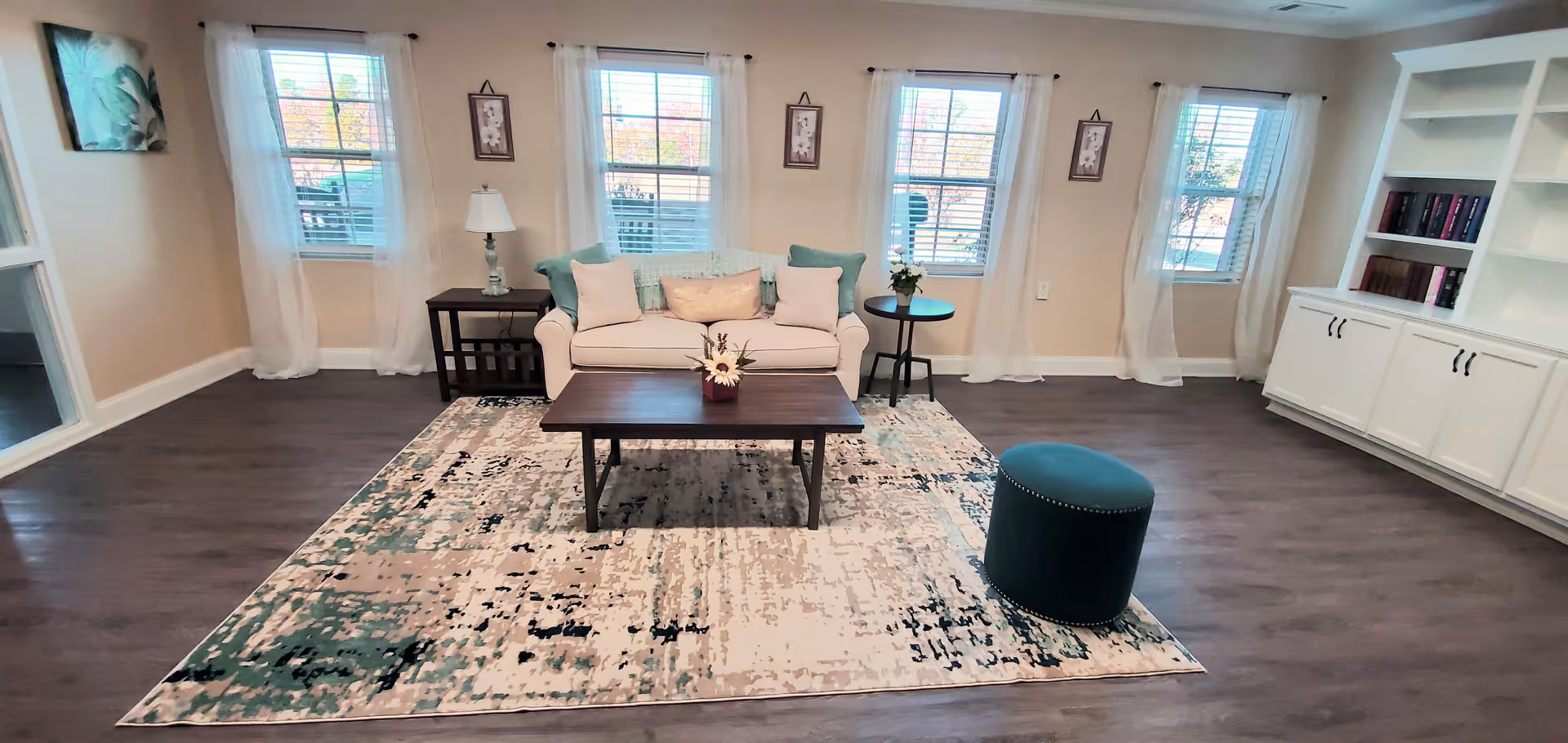 A bright living room with a white sofa adorned with multiple cushions, a wooden coffee table with a small flower arrangement, a teal round ottoman, and two side tables with a lamp and a plant. The room has four windows with white sheer curtains, light beige walls, dark wood flooring, and a large patterned area rug. There is a white built-in bookshelf with books on the right side of the room.