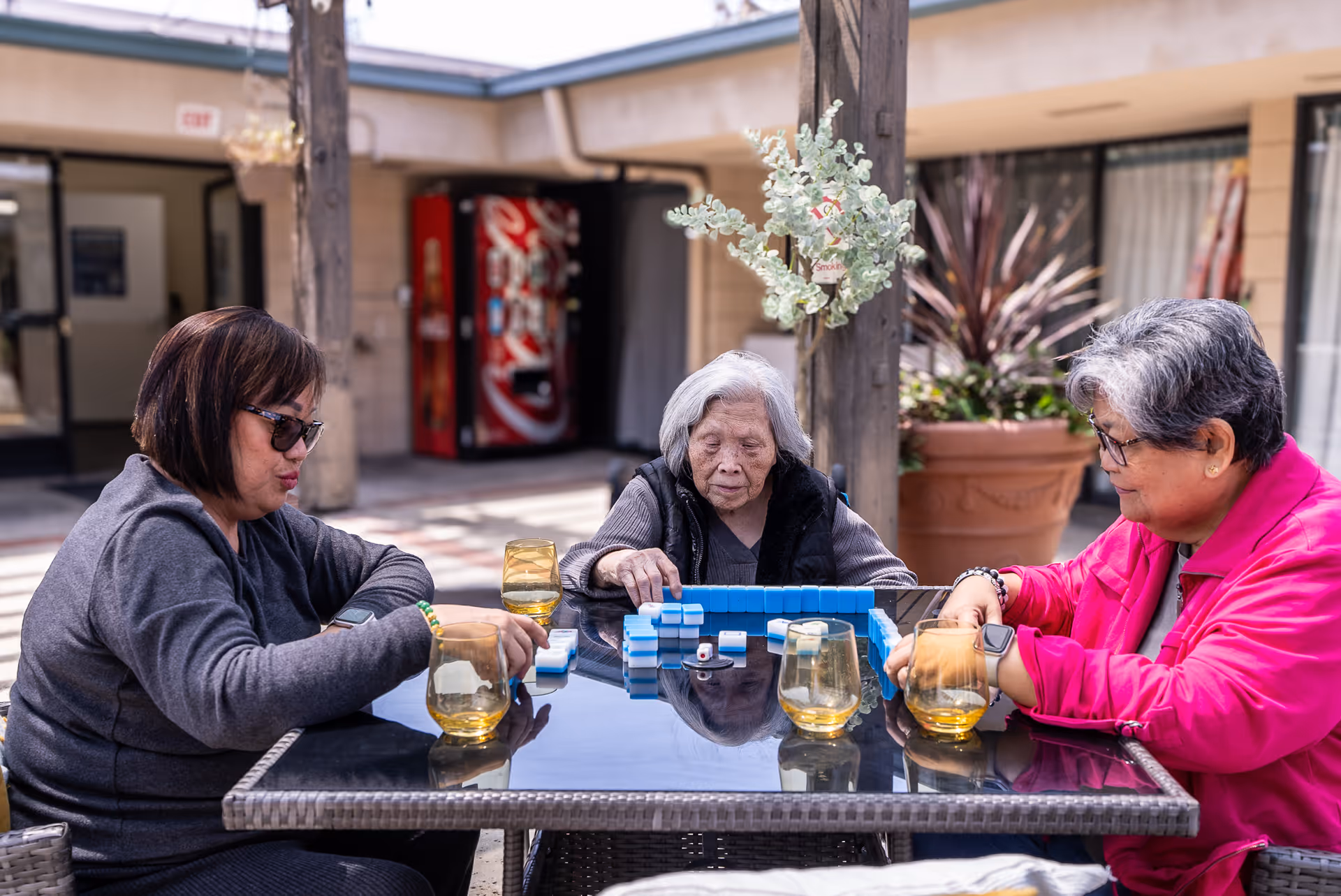 Three elderly women sitting around a glass outdoor table playing a game with blue tiles, with four yellow-tinted glasses on the table. They are in a courtyard area with plants and a vending machine in the background.