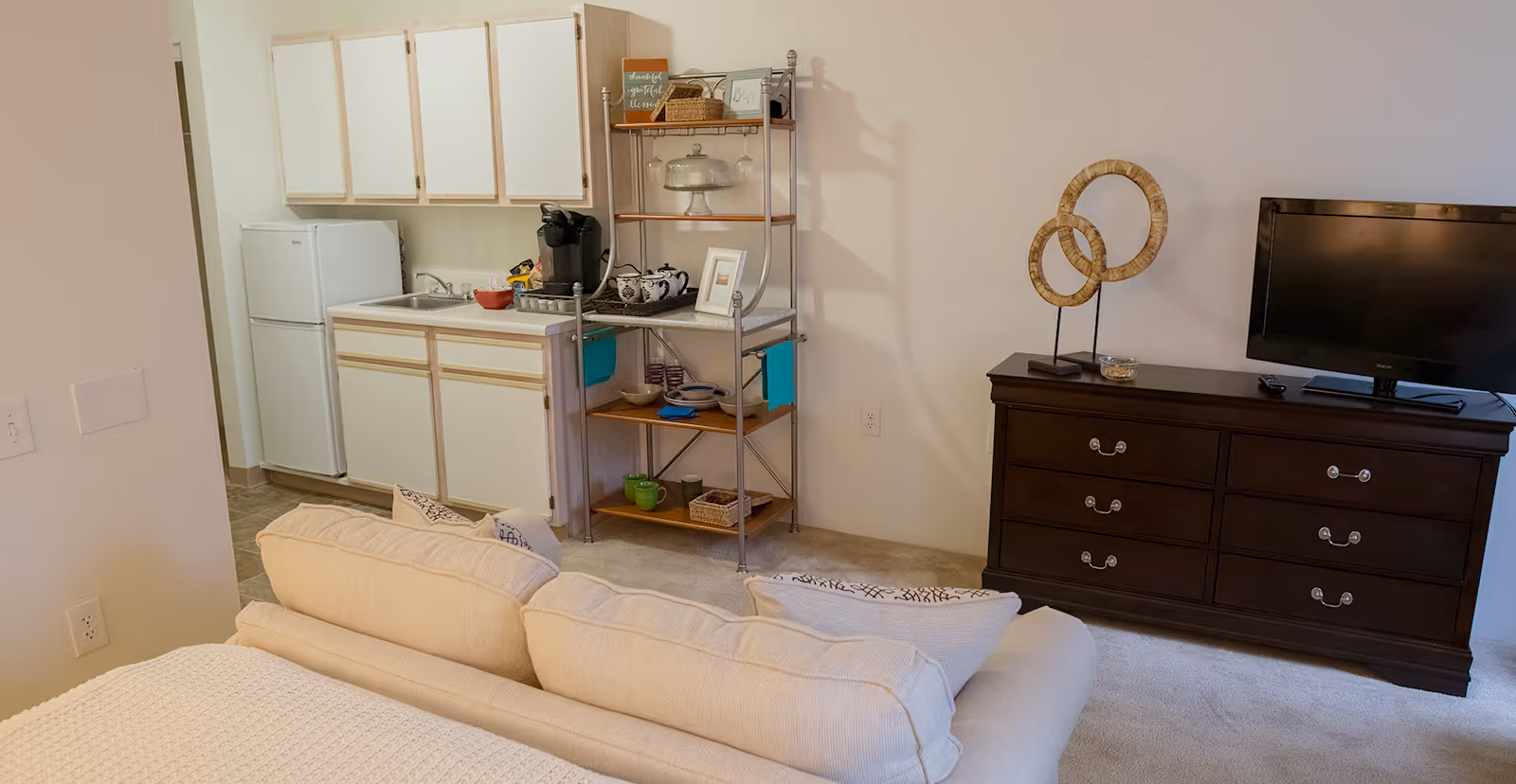 A cozy senior living studio apartment featuring a small kitchenette with white cabinets, a refrigerator, and a sink. Next to the kitchenette is a metal shelving unit holding various kitchen items and decor. In the foreground, there is a beige sofa with pillows, and to the right, a dark wooden dresser with a flat-screen TV and decorative circular sculptures on top.