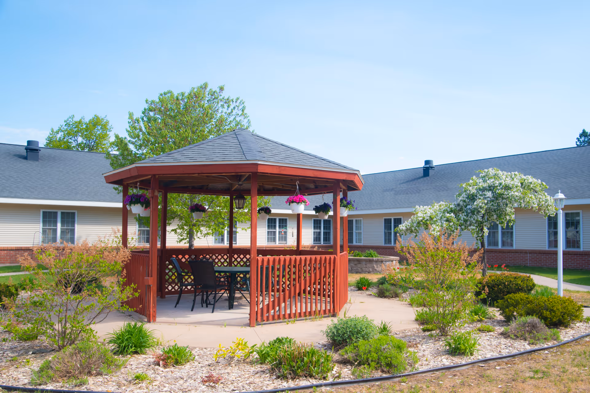 A red wooden gazebo with hanging flower pots and outdoor chairs and table, situated in a landscaped garden area with shrubs and small trees, surrounded by a single-story building with beige siding and a dark roof under a clear blue sky.