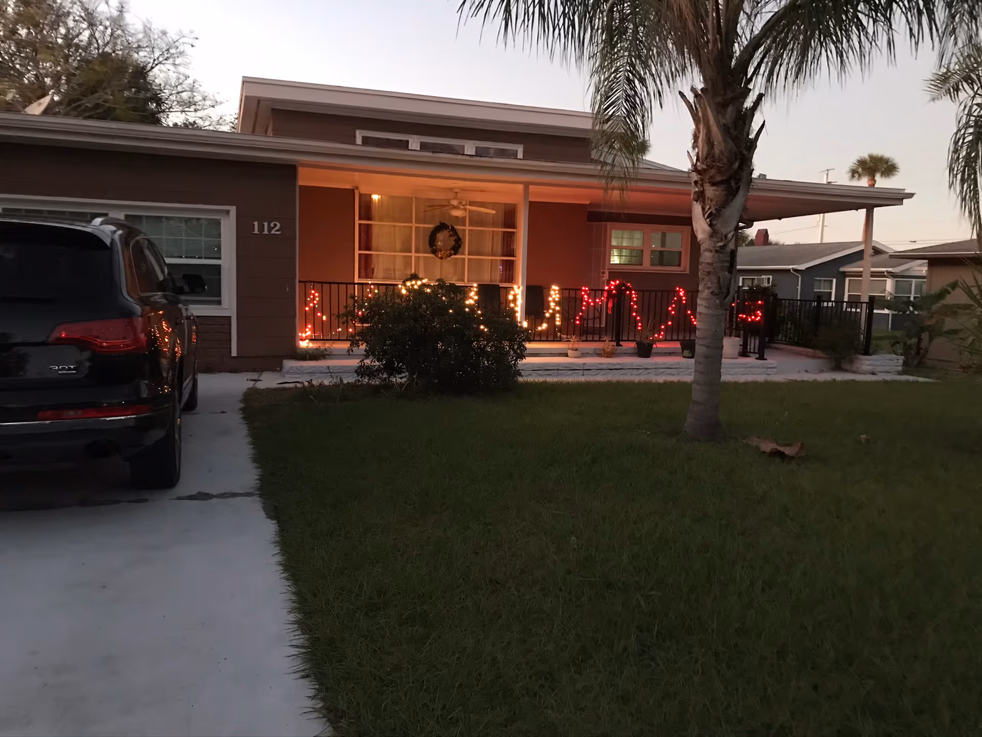 Front exterior view of a single-story house with a driveway and a black SUV parked on it. The house has a covered porch decorated with Christmas lights and a wreath on the window. There is a palm tree and green lawn in the front yard.