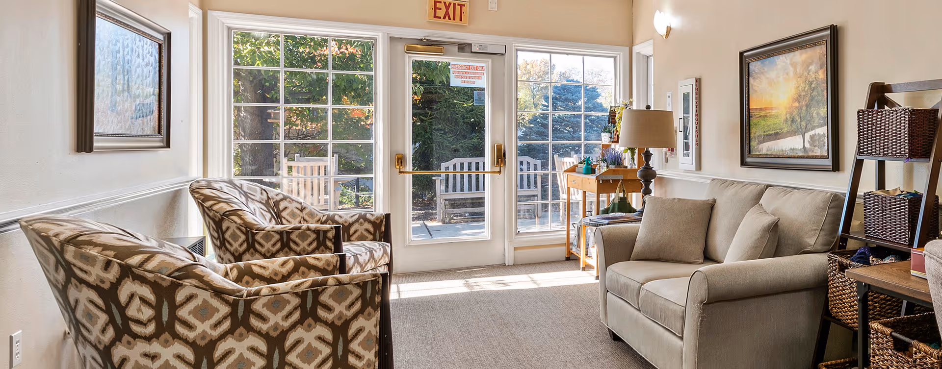Sunlit common sitting area with patterned armchairs, a sofa, side tables and a lamp facing glass exit doors to an outdoor bench.