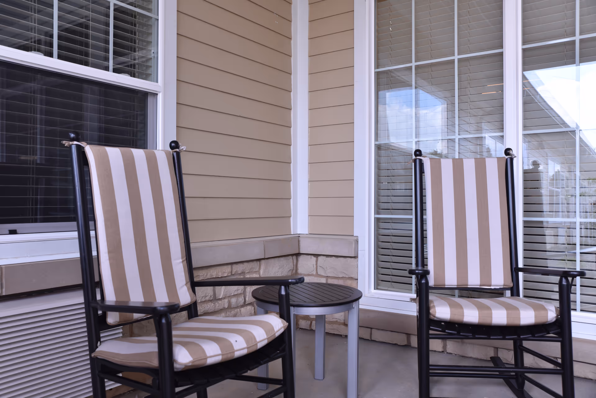 Two striped cushioned chairs and a small round table on a covered patio area with beige siding and large windows with white blinds.