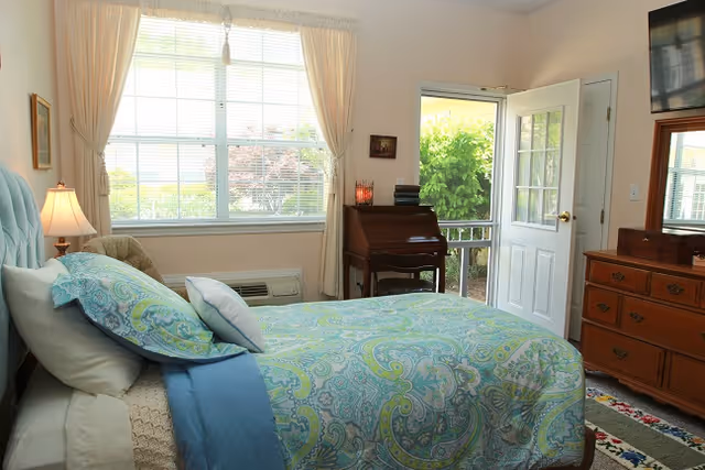 A bright bedroom with a bed covered in blue and green patterned bedding, a window with cream curtains letting in natural light, a wooden dresser with a TV mounted above it, and a door leading outside to a green garden area. There is also a small wooden desk with books and a candle on it.