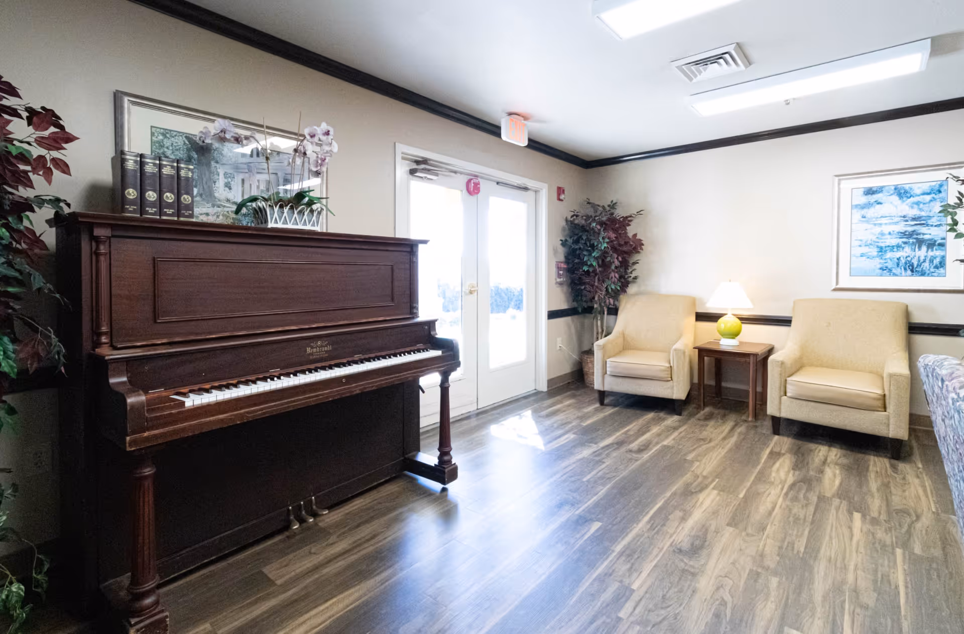 Bright lounge area with an upright piano, two armchairs and a side table with a lamp near double doors.