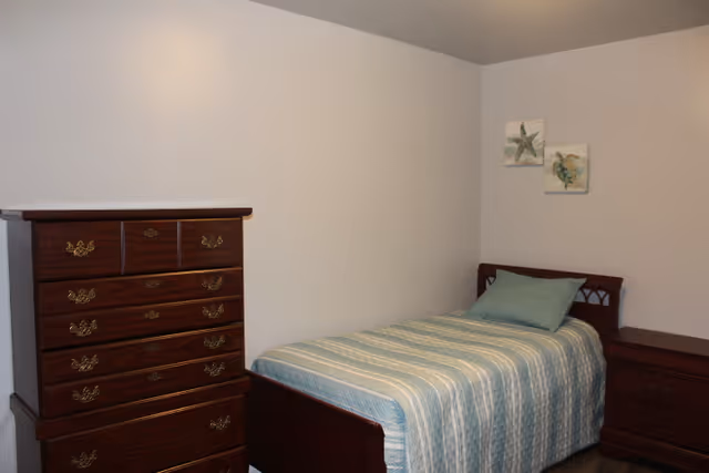 A simple bedroom with a single bed covered in a blue and white striped bedspread and a matching pillow. Next to the bed is a wooden nightstand with two drawers. On the left side of the image is a tall wooden dresser with multiple drawers and brass handles. The walls are plain white with two small framed pictures of sea creatures hanging above the bed.