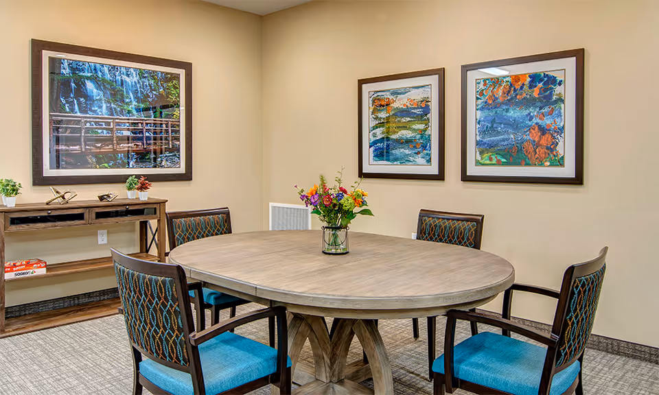 A small meeting or dining room with a round wooden table surrounded by four chairs with blue patterned cushions. A vase with colorful flowers is placed in the center of the table. On the walls, there are three framed artworks: one large photograph of a waterfall and two abstract paintings. A wooden console table with small decorative items and a board game is positioned against the wall.
